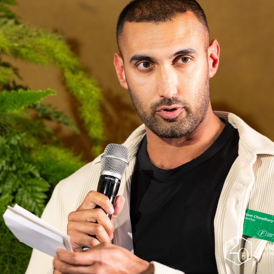 A man in a light jacket holds a microphone and papers, speaking while seated in front of green foliage.